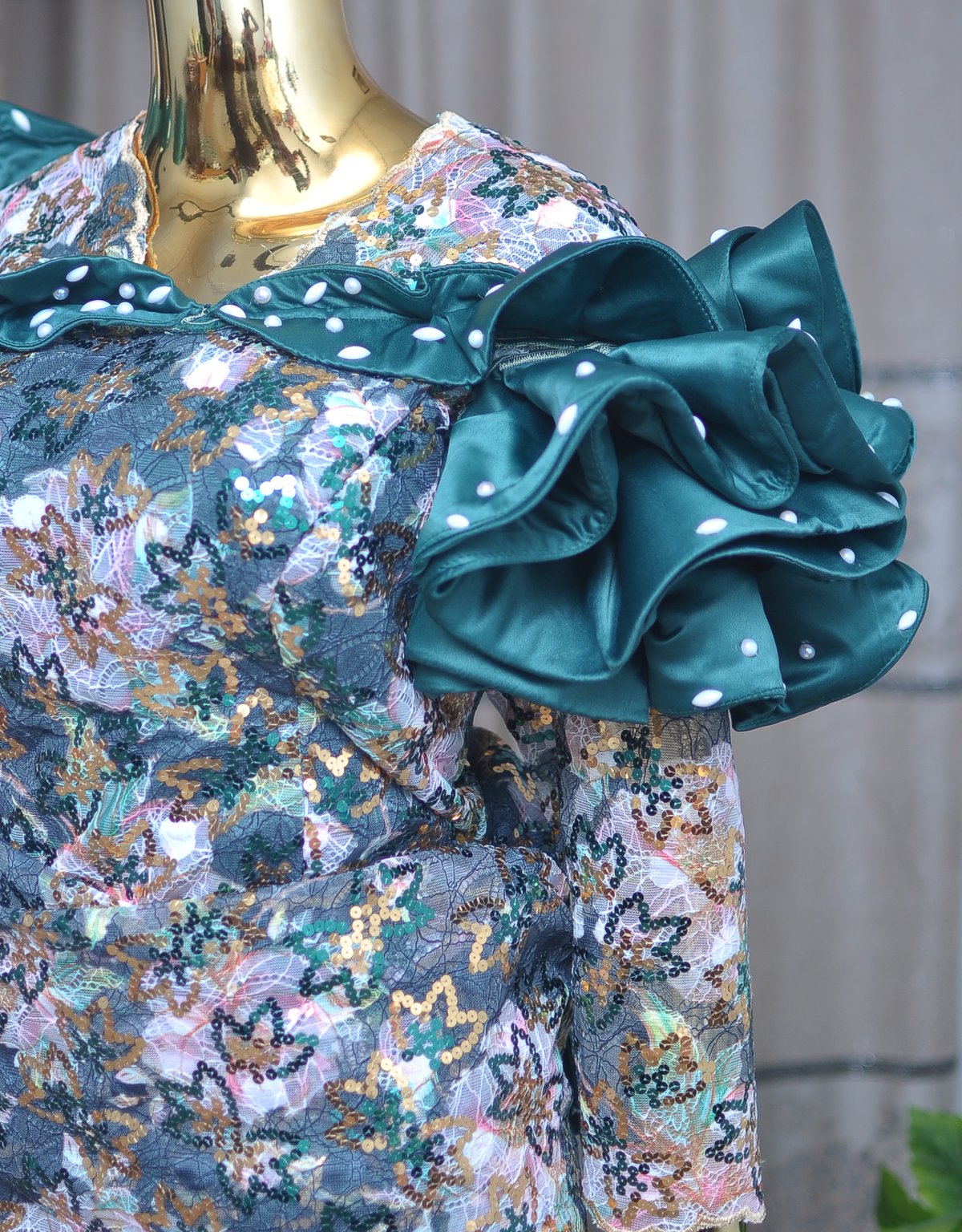 Side view of a sequin lace gown with a sculptural satin ruffle shoulder detail, finished with hand-set pearls.